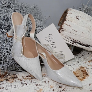 May include: A pair of white, pointed-toe slingback heels with a bow detail and a card that says "Bella". The shoes are embellished with a shimmering, textured material. The background includes white branches and a white-painted log.