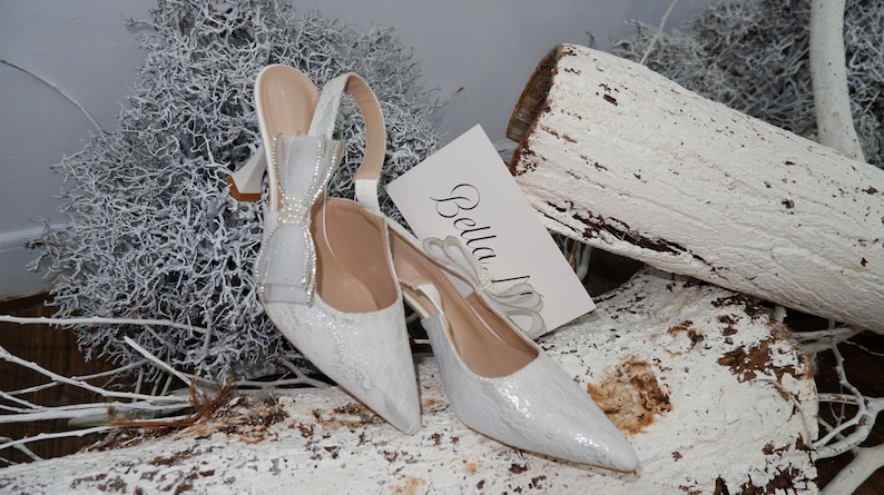 May include: A pair of white, pointed-toe slingback heels with a bow detail and a card that says "Bella". The shoes are embellished with a shimmering, textured material. The background includes white branches and a white-painted log.