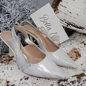 May include: A pair of white, pointed-toe slingback heels with a textured pattern. The heels have a bow and pearl embellishments. A card with the name "Bella" is visible. The shoes are set on a white and brown textured surface.