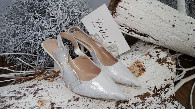 May include: A pair of white, pointed-toe slingback heels with a textured pattern. The heels have a bow and pearl embellishments. A card with the name "Bella" is visible. The shoes are set on a white and brown textured surface.