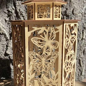 Puede incluir: Linterna de madera con intrincados diseños calados de mariposas y flores. La linterna hexagonal tiene una base y un techo puntiagudo con un asa circular. El color natural de la madera proporciona una estética cálida. Los calados permiten el paso de la luz.