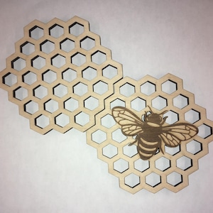 May include: Wooden laser-cut honeycomb-shaped coasters with a detailed bee design. The coasters are made of light-coloured wood and feature a cutout honeycomb pattern. The bee is intricately designed with wings and body details.