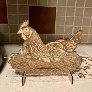 May include: A wooden egg holder in the shape of a hen, featuring a detailed engraving of the bird and its nest. The holder has clear acrylic shelves with circular cutouts for eggs. The design is in shades of brown and beige.