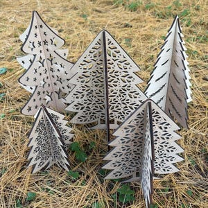 May include: A collection of five freestanding wooden tree sculptures featuring intricate laser-cut designs. The trees display various patterns including star cutouts, leaf motifs, and simple branch silhouettes, all crafted from light-colored wood.