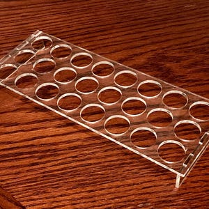 Op de afbeelding: Een heldere acryl displaystandaard met meerdere cirkelvormige openingen. De rechthoekige standaard heeft een opstaande rand aan één kant en is ontworpen om kleine items in te plaatsen. De standaard staat op een houten oppervlak.