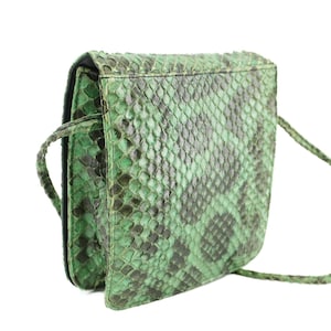 May include: A small, square, green snakeskin crossbody bag. The bag features a flap closure and a long, thin strap. The snakeskin pattern is a mix of green and black, giving it a textured appearance. The bag is shown against a white background.