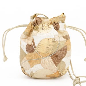 May include: A drawstring pouch bag with a patchwork design. The bag features a variety of neutral tones, including beige, cream, and brown, with a mix of textures. The bag has a drawstring closure and a cream-colored cord.