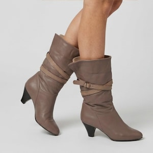 May include: A pair of taupe mid-calf boots featuring a slouchy shaft design, a pointed toe, and a black kitten heel. Each boot is accented with a thin, wrap-around strap detail that features a small buckle closure near the top of the shaft.