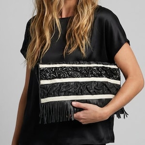 May include: Black clutch with white stripes and fringe. The clutch has a textured design and is held by a person wearing a black top. The bag has a rectangular shape and a decorative pattern.