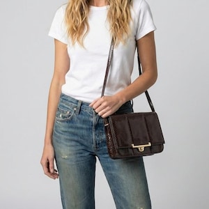 May include: A dark brown crossbody bag with a rectangular shape and a gold-colored clasp. The bag has a textured surface and a long, thin strap. The bag is worn over the shoulder.
