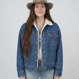 May include: A blue denim jacket with a white sherpa lining, featuring a button-up front, two chest pockets, and long sleeves. The jacket is paired with a light brown felt fedora hat with a thin leather band and a turquoise beaded necklace.