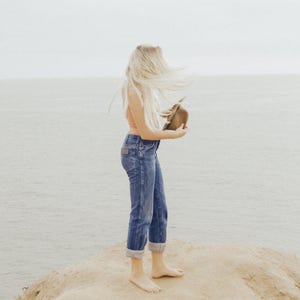 Op de afbeelding: Een vrouw met lang blond haar staat op een zanderige klif met uitzicht op de oceaan. Ze draagt een lichtroze tanktop en een blauwe jeans. Ze houdt een bruine hoed in haar hand.
