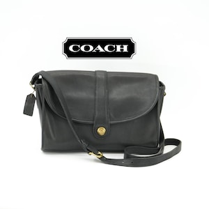 May include: Black leather Coach crossbody bag with a flap closure and adjustable strap. The bag features a gold-tone clasp and hardware. A small leather tag hangs from the side. The Coach logo is displayed at the top.