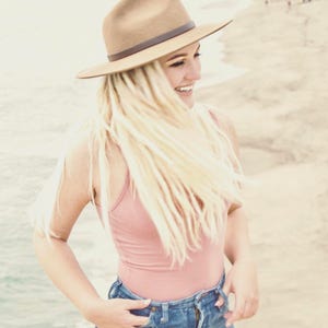 Op de afbeelding: Een vrouw met lang blond haar die een bruine fedora, een roze tanktop en een blauwe jeans draagt. Ze glimlacht en kijkt naar de zijkant.