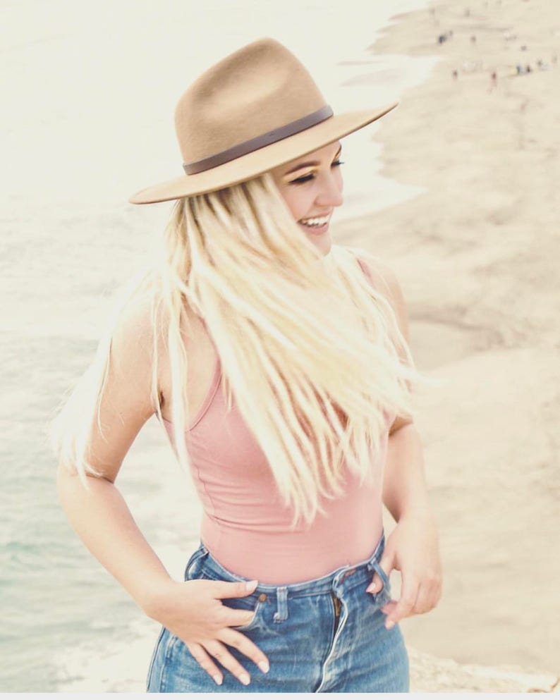 Op de afbeelding: Een vrouw met lang blond haar die een bruine fedora, een roze tanktop en een blauwe jeans draagt. Ze glimlacht en kijkt naar de zijkant.