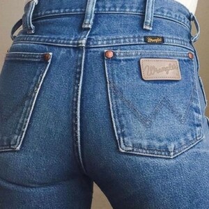 Op de afbeelding: Een paar blauwe denim jeans met een achterzak met een bruin leren patch met het woord "Wrangler" in witte letters.