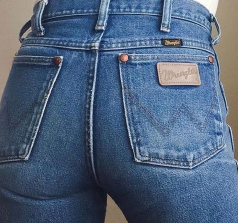 Op de afbeelding: Een paar blauwe denim jeans met een achterzak met een bruin leren patch met het woord "Wrangler" in witte letters.