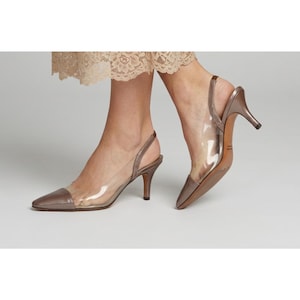 May include: A pair of slingback heels featuring a pointed toe and a mid-height stiletto heel. The shoes are constructed with a metallic bronze-toned leather toe cap and heel counter, contrasted by transparent clear side panels along the foot.