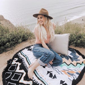 Op de afbeelding: Een vrouw met een bruine hoed, een roze tanktop en een blauwe jeans zit op een ronde zwart-witte strandhanddoek met een geometrisch patroon. De handdoek heeft een franje rand.