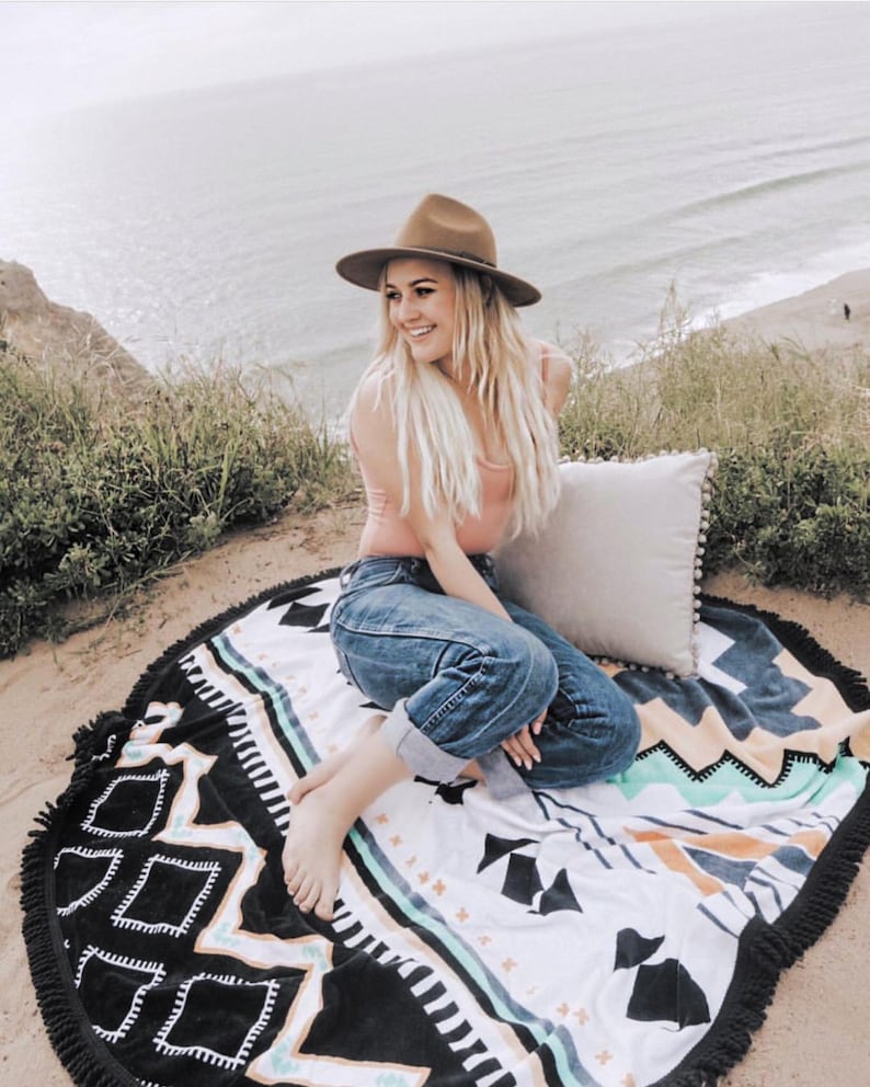 Op de afbeelding: Een vrouw met een bruine hoed, een roze tanktop en een blauwe jeans zit op een ronde zwart-witte strandhanddoek met een geometrisch patroon. De handdoek heeft een franje rand.