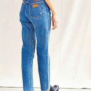 Op de afbeelding: Een persoon draagt ​​een paar high-waisted blauwe denim jeans met een lichtbruine leren patch op de achterzak.