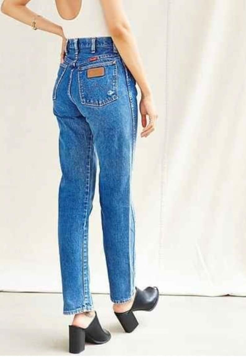Op de afbeelding: Een persoon draagt ​​een paar high-waisted blauwe denim jeans met een lichtbruine leren patch op de achterzak.