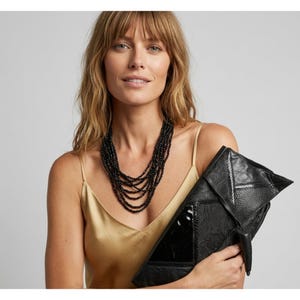 May include: A woman wearing a multi-strand black beaded necklace and a gold satin camisole. She is holding a black leather clutch with a geometric design and a glossy panel. The clutch has a textured, embossed pattern.