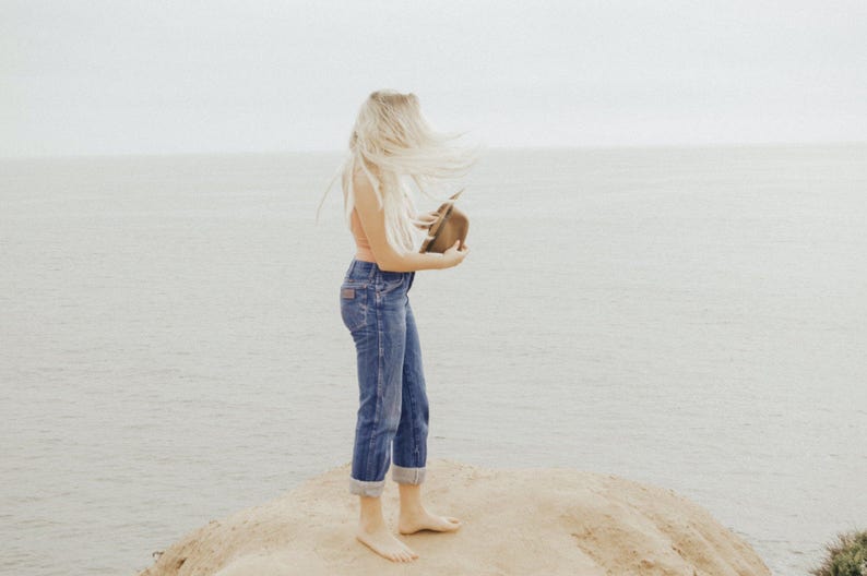 Op de afbeelding: Een vrouw met lang blond haar staat op een zanderige klif met uitzicht op de oceaan. Ze draagt een lichtroze tanktop en een blauwe jeans. Ze houdt een bruine hoed in haar hand.