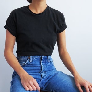 Op de afbeelding: Een vrouw draagt een zwart T-shirt en een blauwe jeans. Ze draagt een hoed en zit op een houten kruk.