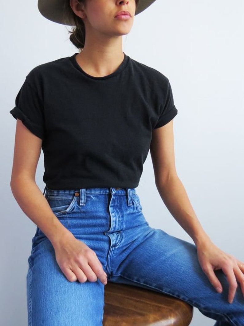 Op de afbeelding: Een vrouw draagt een zwart T-shirt en een blauwe jeans. Ze draagt een hoed en zit op een houten kruk.