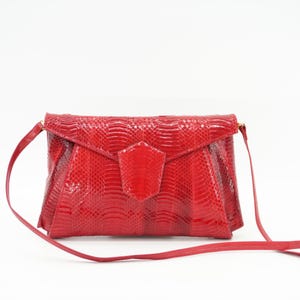 May include: A red, textured clutch bag with a long, thin shoulder strap. The bag features a flap closure and a geometric design. The material has a snakeskin pattern. The bag is a vibrant shade of red.