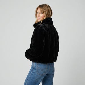 May include: A black faux fur jacket with a high collar. The jacket is cropped and has long sleeves. The model is wearing blue jeans. The background is a solid light gray.