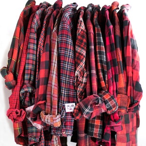 May include: A collection of red and black plaid flannel shirts hanging on a rack. The shirts have rolled-up sleeves and various plaid patterns, including classic buffalo check and tartan designs. Some shirts have tags attached.