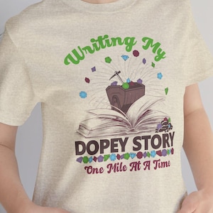 May include: A heather beige t-shirt featuring a graphic of an open book with a mining cart and pickaxe on top. The text reads "Writing My DOPEY STORY One Mile At A Time" in green and maroon, surrounded by colourful gemstone illustrations.
