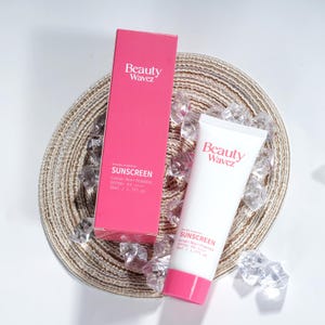 May include: Pink and white sunscreen tube with the text "Beauty Wavez" and "SUNSCREEN" on the front. The tube is sitting on a woven mat with ice cubes. The product is also available in a pink box with the same text.
