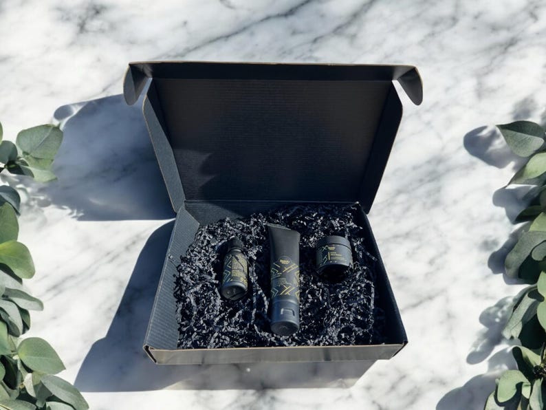 Beauty Wavez skincare gift set with cleanser, vitamin C serum, and facial shea butter in a premium black box with gold accents, styled on a luxury white marble background with eucalyptus sprigs.