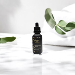 Beauty Wavez Vitamin C Serum in matte black dropper bottle, staged on clean marble with spa towel and eucalyptus leaves, ideal for daily brightening skincare.