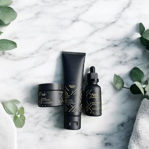 Beauty Wavez daily skincare trio arranged on a spa-style white marble counter with folded towels and eucalyptus leaves, featuring sleek matte black packaging with gold geometric design.