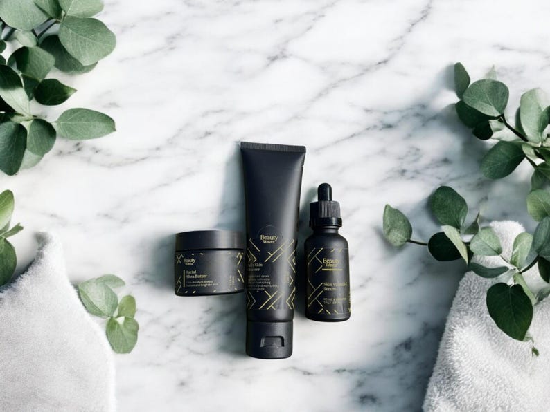 Beauty Wavez daily skincare trio arranged on a spa-style white marble counter with folded towels and eucalyptus leaves, featuring sleek matte black packaging with gold geometric design.