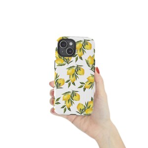 May include: A white phone case with a pattern of yellow lemons and green leaves. The case is held in a hand against a white background. The phone case has a black camera lens.