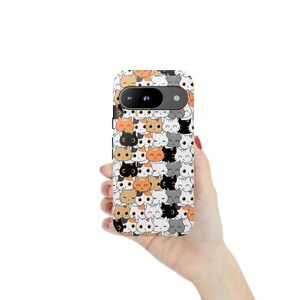 Puede incluir: Una funda de teléfono blanca cubierta con un patrón repetido de caras de gatos de dibujos animados en varios colores, incluyendo naranja, negro, gris y blanco. La funda del teléfono tiene una lente de cámara y un flash negros.