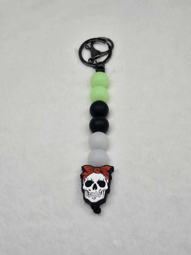 May include: A black and white skull keychain with a red bandana. The keychain has a silver metal ring and is made of silicone beads in green, black, white, and gray.