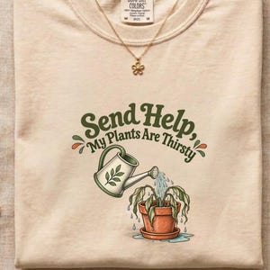 Peut inclure: Un t-shirt beige à col rond avec un motif représentant un arrosoir vert versant de l'eau sur une plante en pot. Le texte "Send Help, My Plants Are Thirsty" est écrit en vert foncé. Un collier pendentif fleur dorée repose sur le col.