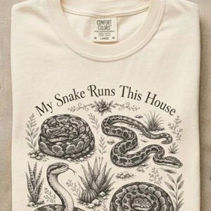 Op de afbeelding: Een crèmekleurig t-shirt met een zwarte inktillustratie van drie slangen en diverse planten. De tekst "My Snake Runs This House" is in een boog boven de slangen gedrukt. Het t-shirt heeft een ronde hals en een Comfort Colors label.
