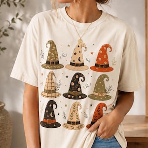 Peut inclure: Un t-shirt à manches courtes de couleur crème arborant un imprimé graphique composé de neuf chapeaux de sorcière distincts disposés en grille. Les chapeaux présentent divers motifs d'étoiles, de lunes, de fleurs et de carreaux en vert, orange et noir.
