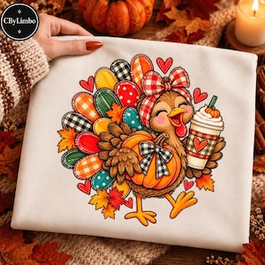 Peut inclure: Un t-shirt blanc avec un motif coloré représentant une dinde portant un nœud à carreaux rouge et blanc, tenant une citrouille et une boisson à la crème fouettée. La dinde a des plumes aux motifs variés, dont des pois et des carreaux.