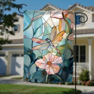 May include: A decorative garden flag featuring a stained-glass design. The flag showcases a dragonfly with orange and red wings, a light blue body, and a pink and white flower. The background is a mix of blue, green, and peach tones.