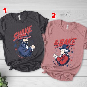May include: A dark gray t-shirt with the text SHAKE and a graphic of a man in a red hat, and a dusty rose t-shirt with the text & BAKE and a graphic of a man in a blue hat. Both shirts feature red and blue star accents and historical attire.