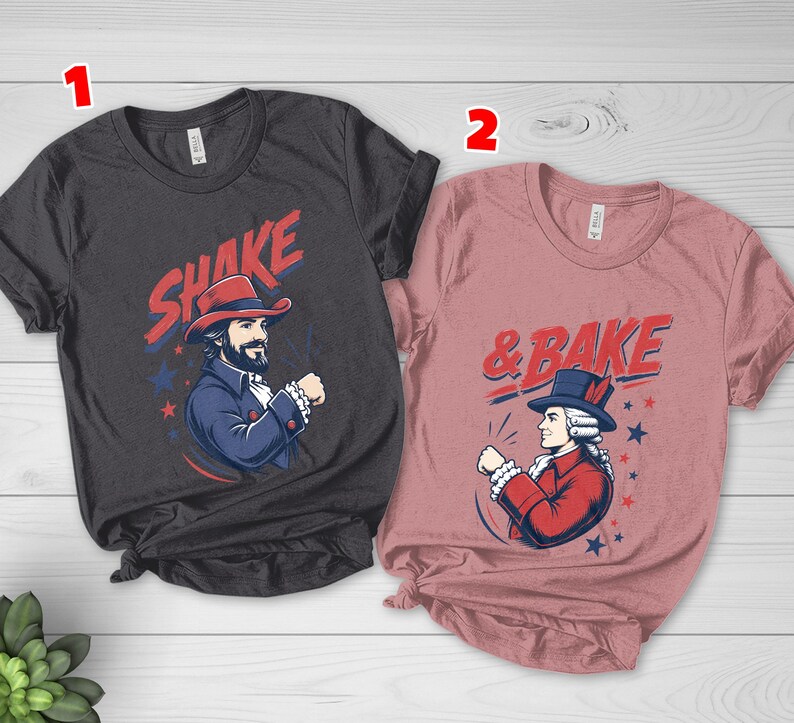 May include: A dark gray t-shirt with the text SHAKE and a graphic of a man in a red hat, and a dusty rose t-shirt with the text & BAKE and a graphic of a man in a blue hat. Both shirts feature red and blue star accents and historical attire.
