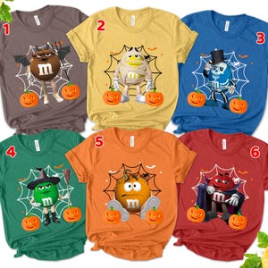 May include: A collection of six Halloween-themed t-shirts. Each shirt features a different M&M's character dressed in a Halloween costume, such as a mummy, skeleton, and witch. Each design includes a spider web and pumpkins.